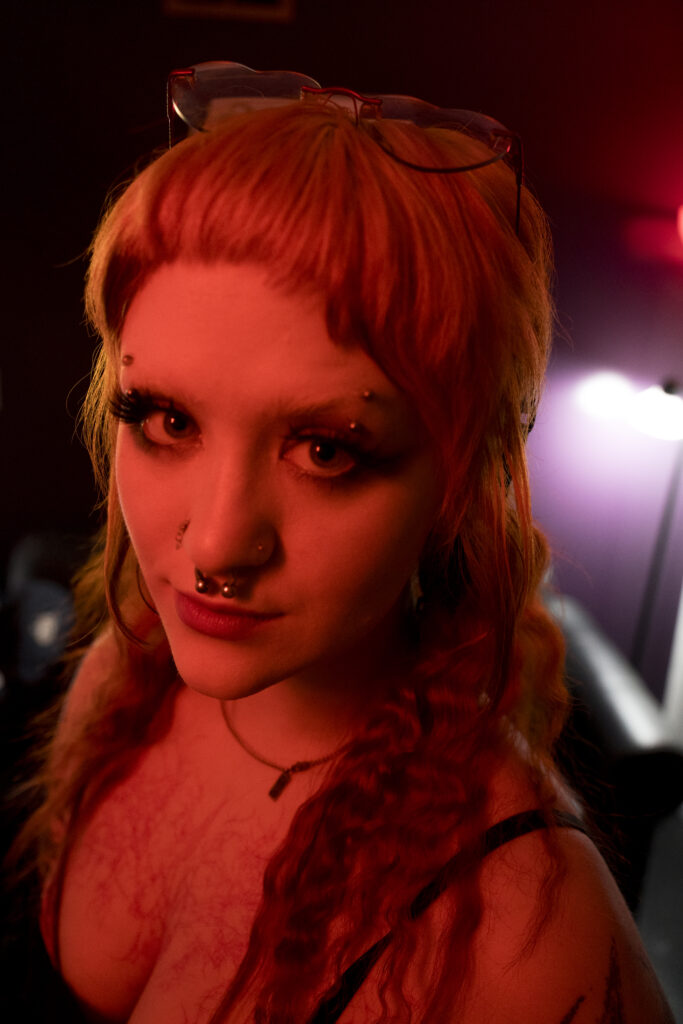 A person looks directly at viewer, they have various piercings and tattoos.